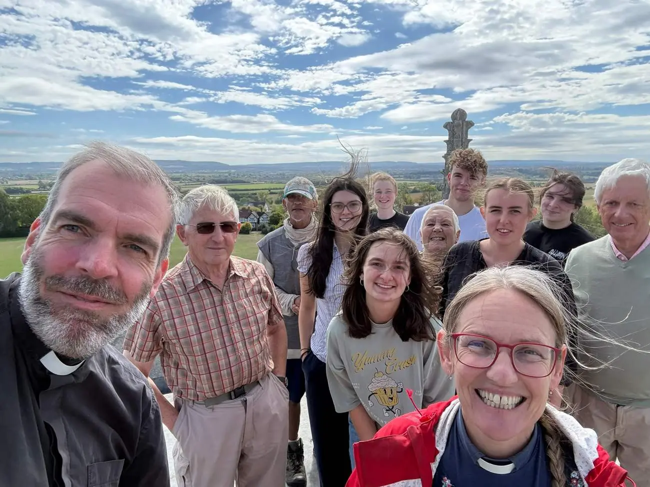 A group of church members of all ages smiling together on the roof of a church tower, with fields and hills stretching out behind them.